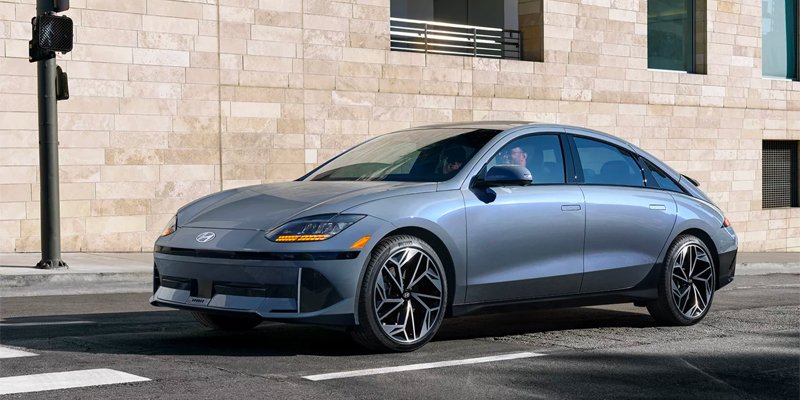 A sleek gray Hyundai 2025 Hyundai IONIQ 6 electric car parked on the street with a modern building in the background.