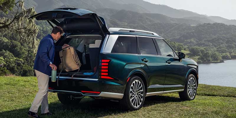 A man loading luggage into the rear of a green 2026 Hyundai Palisade Hybrid in a scenic outdoor setting.